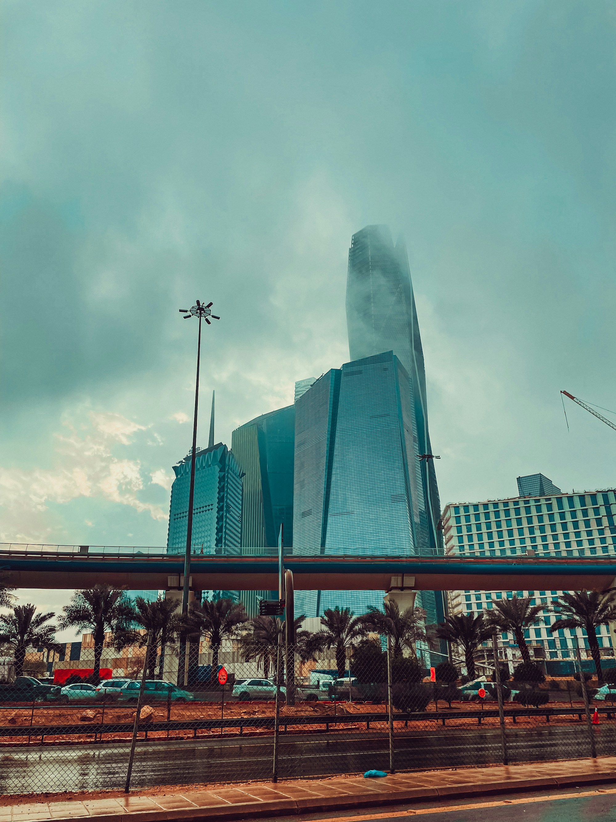 Riyadh towers above a road and palm trees under a cloudy sky